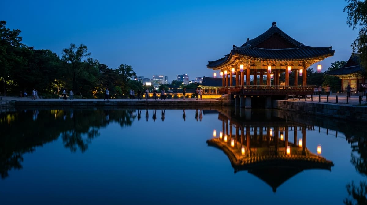 Evening view of a palace pavilion reflected in a still pond, warm lanterns glowing at dusk and silhouettes of visitors on the far bank, no signage