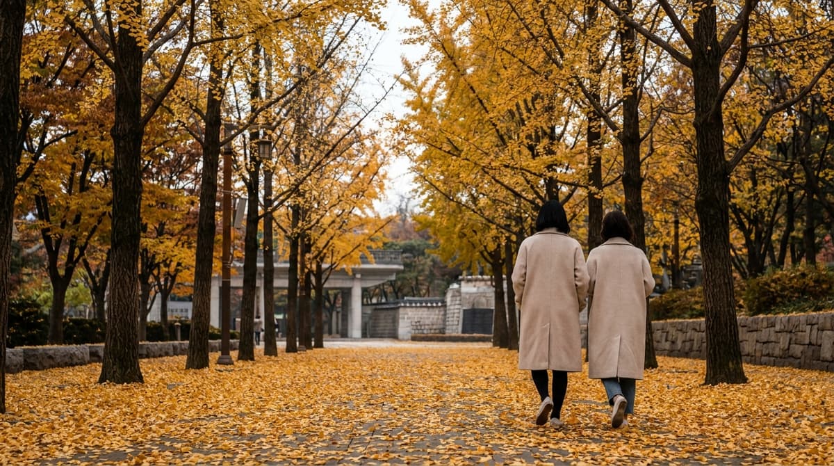 A broad city park walkway in late autumn, thick carpet of fallen yellow ginkgo leaves underfoot, tall trees arching overhead in gold and amber, two people in light coats walking away from the camera, soft overcast afternoon light, no signs or text visible