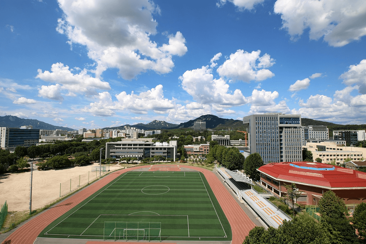 High-angle panoramic view of a university campus featuring a green soccer field with a running track under a bright blue sky with fluffy white clouds.