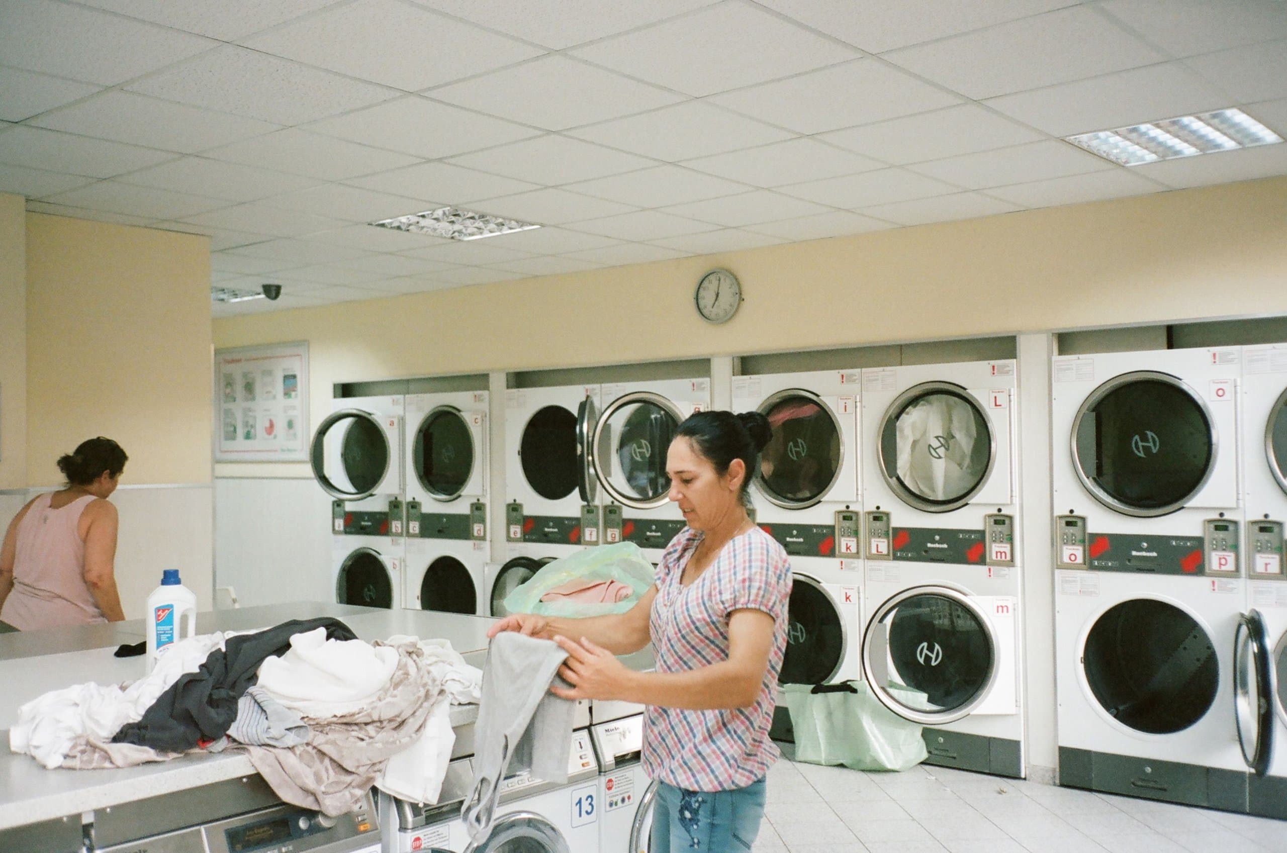 A woman folding laundry inside a self-service laundromat with rows of washing machines and dryers in the background.