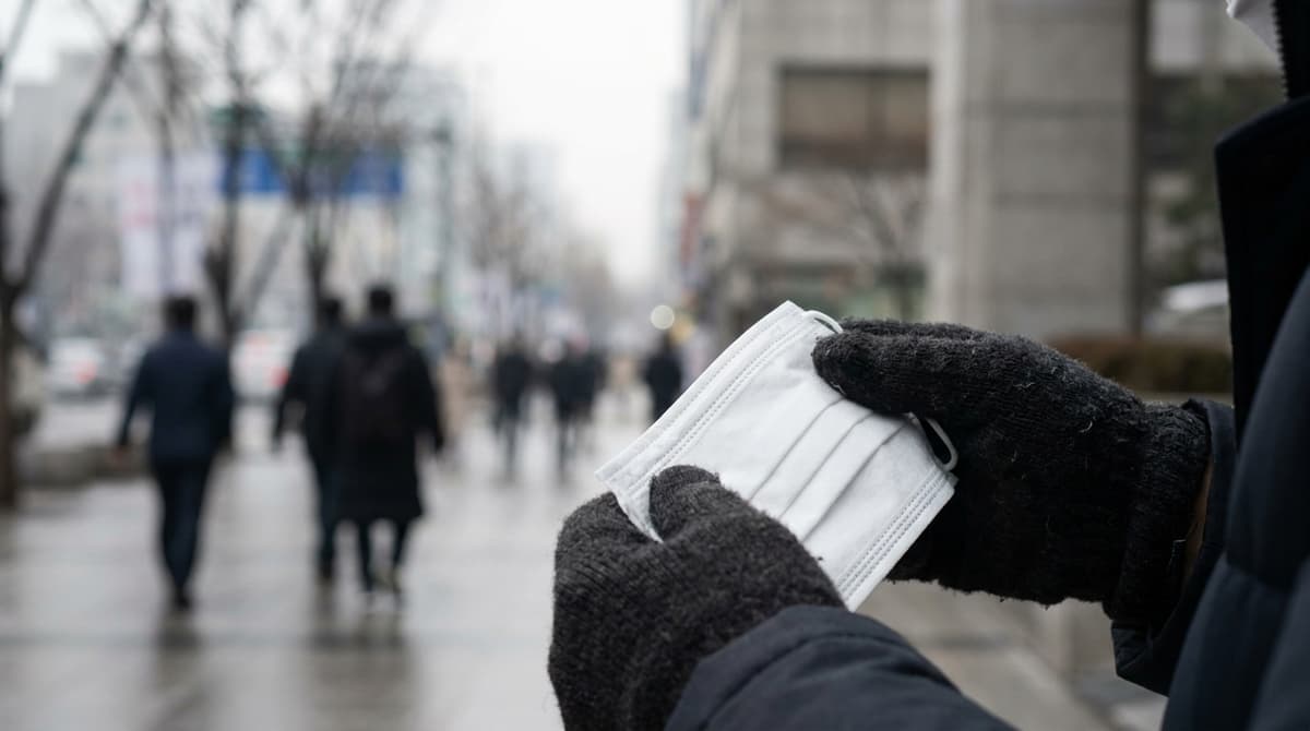 Close-up of a person's gloved hands holding a completely plain white folded disposable face mask with no markings, outdoors on a hazy grey winter day, a blurred city street and bare trees behind, shallow depth of field, no logos, no printing, no text anywhere