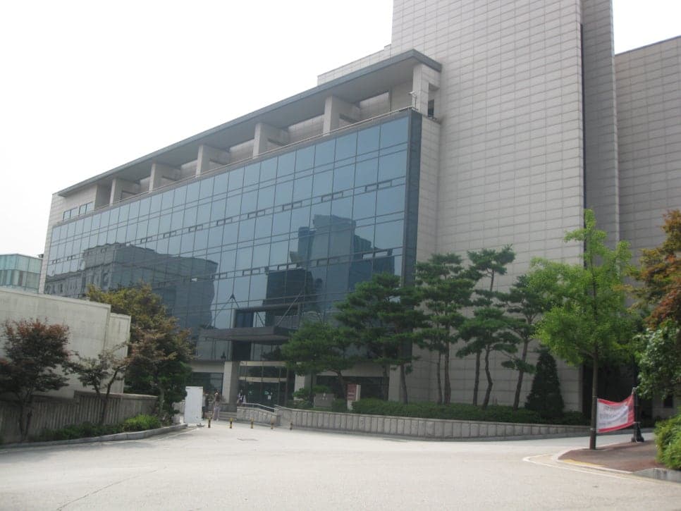 유리 외벽과 밝은 석재로 마감한 캠퍼스 건물 앞에 나무가 늘어서 있습니다.