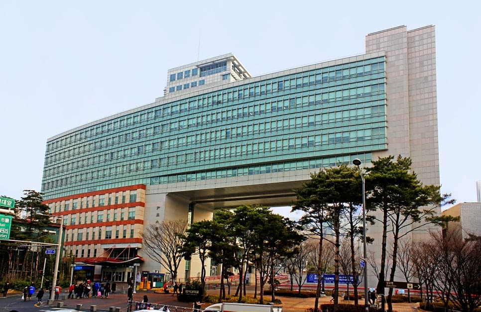 유리창이 길게 이어진 대형 건물 앞에 나무와 보행자가 보인다.