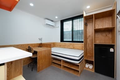 창 아래 매트리스와 왼쪽 책상, 오른쪽 목재 수납장과 냉장고가 놓여 있다.