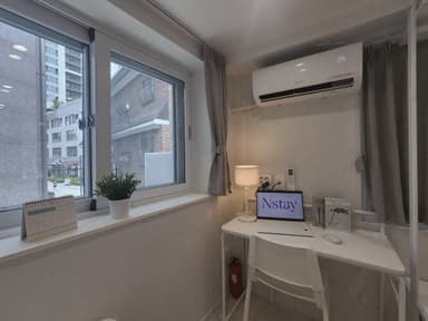창가 책상에 조명과 노트북이 놓여 있고 위쪽 벽에는 에어컨이 설치되어 있다.