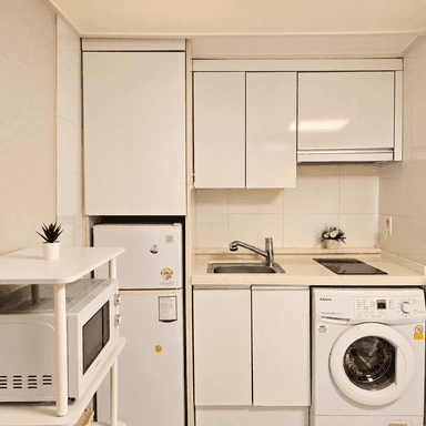 흰색 수납장과 싱크대 아래에 세탁기가 있고 옆에는 냉장고와 전자레인지가 놓여 있다.
