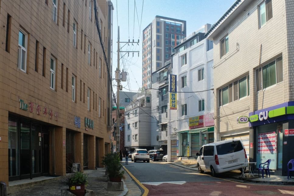 저층 건물이 늘어선 골목에 편의점과 주차된 차량들이 보인다.