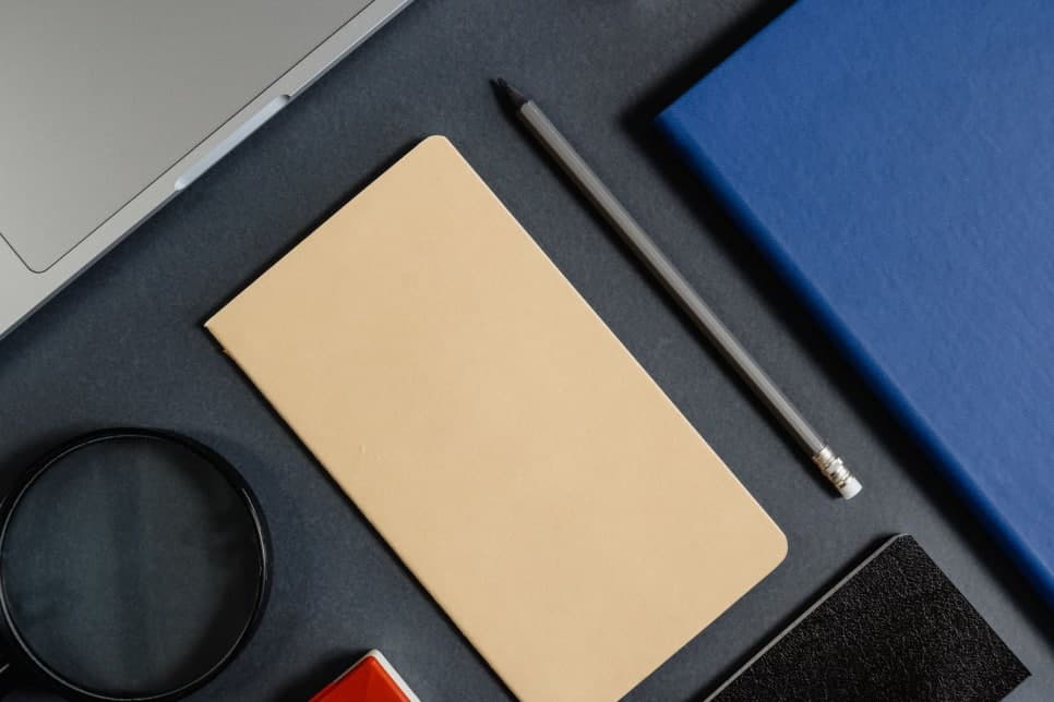 짙은 책상 위에 빈 노트와 연필이 놓이고 노트북 일부가 보인다.