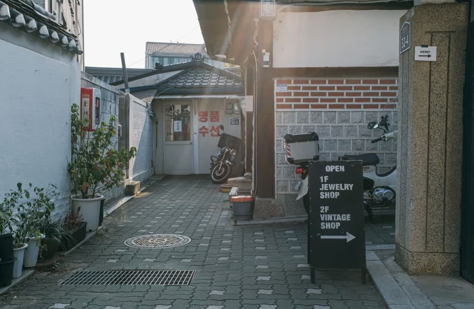 화분이 놓인 좁은 골목에 상점 안내판과 오토바이가 서 있다.