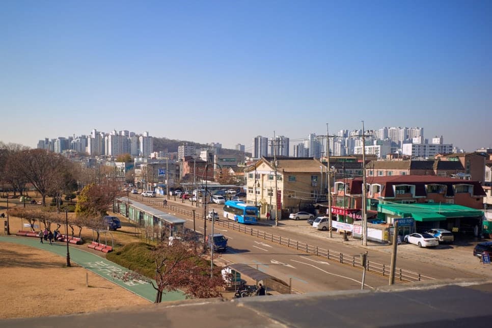 공원 옆 도로에 파란 버스가 지나고 뒤로 주택과 아파트가 보인다.