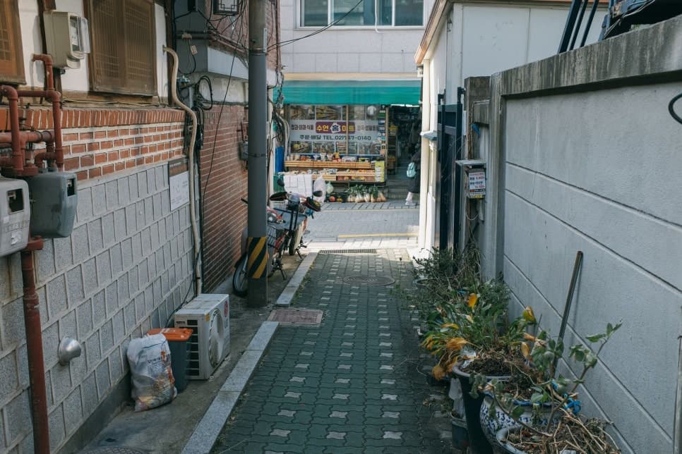 벽돌집과 회색 담장 사이 좁은 골목 끝에 작은 상점이 보인다.