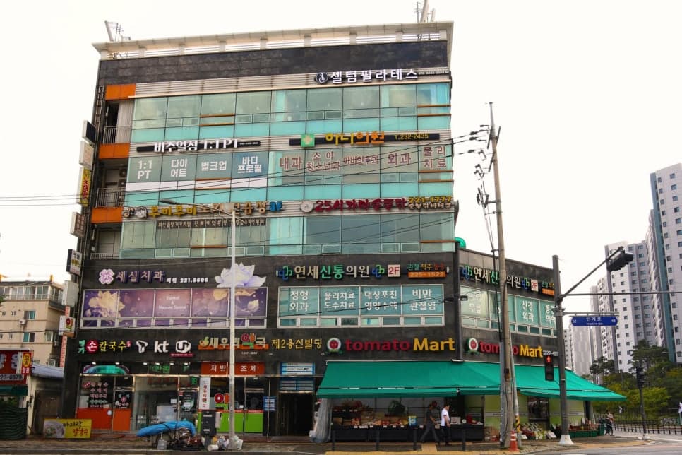 마트와 약국, 여러 병원 간판이 모인 상가 건물이 도로변에 서 있습니다.